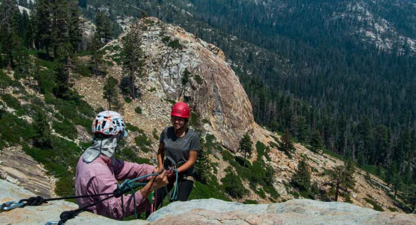 Two people wearing safety gear are secured by ropes near the edge of a cliff. There is a forrest of evergreen trees below. 